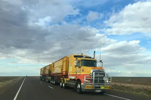 Yellow semi traveling down a road with the sky and clouds in the background.