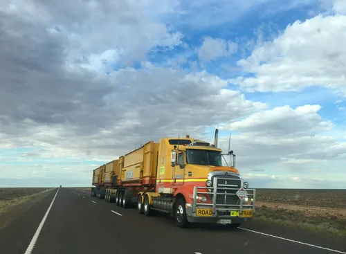 Yellow semi traveling down a road with the sky and clouds in the background.